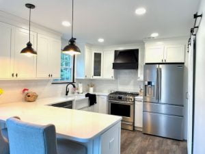 a modern kitchen with white cabinets, black countertops, and stainless steel appliances. The kitchen has a large peninsula with a sink and a faucet, and three stools for seating. The walls are painted in a light gray color, and the floor is made of dark wood. The kitchen has a lot of natural light coming from the windows and the sliding glass door that leads to the backyard. The kitchen is spacious, clean, and elegant.