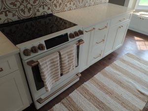 A modern kitchen featuring a white oven with two hanging towels, adjacent to elegant white cabinetry with gold handles, set against a patterned backsplash. The floor is adorned with a striped rug.