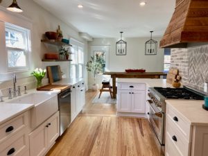 A bright, modern kitchen with white cabinetry, a wooden floor, and a peninsula. The room is adorned with hanging pendant lights and features a herringbone backsplash.